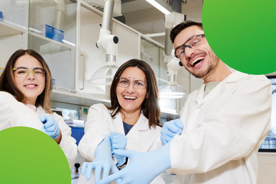 Drei Personen schauen lachend in die Kamera und ziehen sich Gummihandschuhe an. Sie tragen Laborkittel und Schutzbrillen.