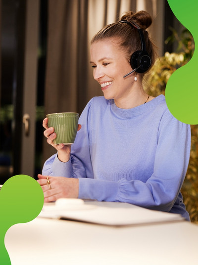 Eine junge Frau sitzt an einem Laptop. Sie lächelt und hält eine Tasse in der Hand. Sie trägt ein Headset und schaut auf den Bildschirm des Laptops.