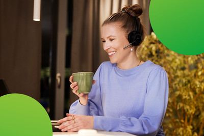 Eine junge Frau sitzt an einem Laptop. Sie lächelt und hält eine Tasse in der Hand. Sie trägt ein Headset und schaut auf den Bildschirm des Laptops.