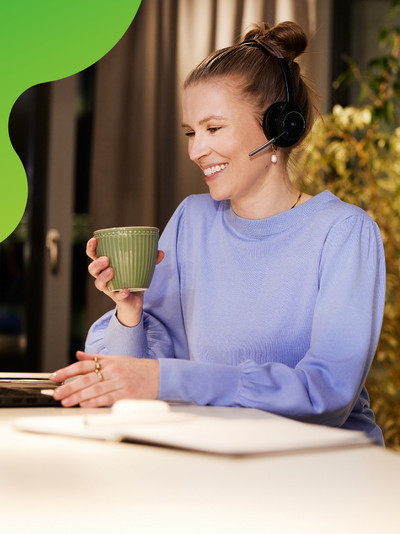 Eine junge Frau sitzt an einem Laptop. Sie lächelt und hält eine Tasse in der Hand. Sie trägt ein Headset und schaut auf den Bildschirm des Laptops.