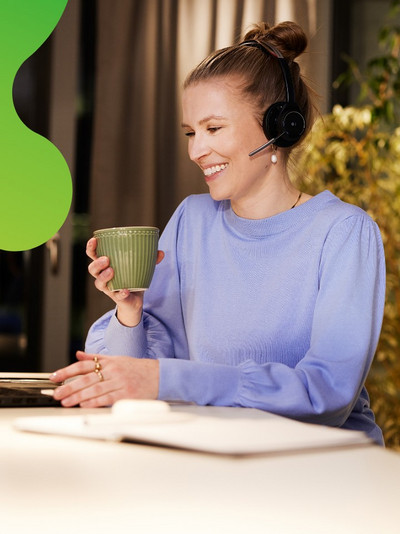Eine junge Frau sitzt an einem Laptop. Sie lächelt und hält eine Tasse in der Hand. Sie trägt ein Headset und schaut auf den Bildschirm des Laptops.