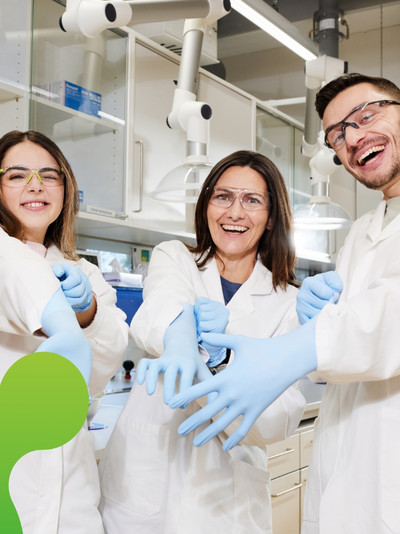 Drei Personen schauen lachend in die Kamera und ziehen sich Gummihandschuhe an. Sie tragen Laborkittel und Schutzbrillen.