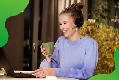Eine junge Frau sitzt an einem Laptop. Sie lächelt und hält eine Tasse in der Hand. Sie trägt ein Headset und schaut auf den Bildschirm des Laptops.