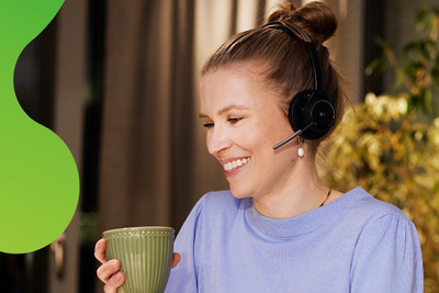 Eine junge Frau sitzt an einem Laptop. Sie lächelt und hält eine Tasse in der Hand. Sie trägt ein Headset und schaut auf den Bildschirm des Laptops.