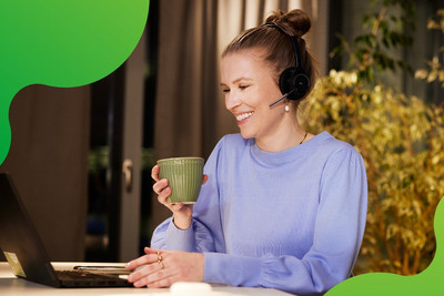 Eine junge Frau sitzt an einem Laptop. Sie lächelt und hält eine Tasse in der Hand. Sie trägt ein Headset und schaut auf den Bildschirm des Laptops.