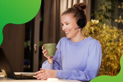 Eine junge Frau sitzt an einem Laptop. Sie lächelt und hält eine Tasse in der Hand. Sie trägt ein Headset und schaut auf den Bildschirm des Laptops.