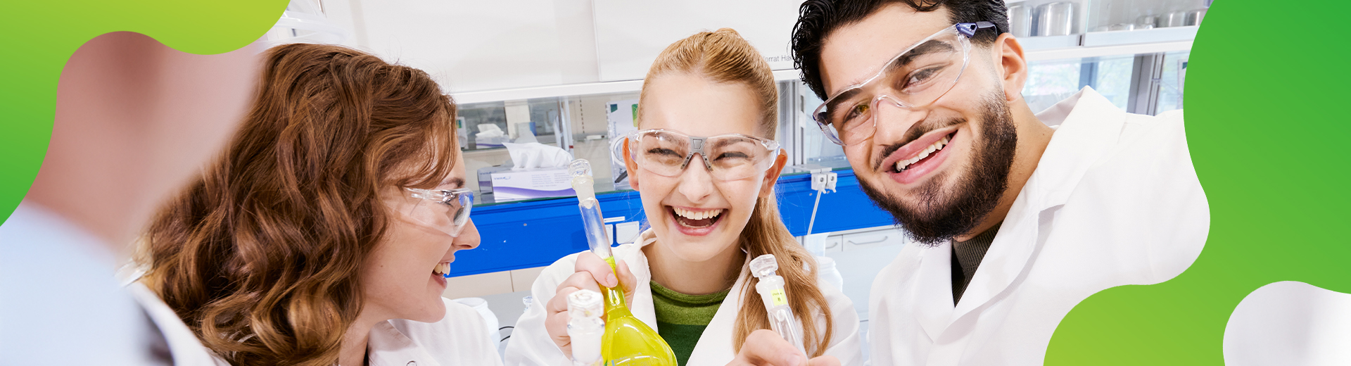 Drei junge Menschen tragen Schutzbrillen und Laborkittel. Eine junge Frau hält Gefäße mit farbigen Flüssigkeiten in der Hand. Alle lachen 