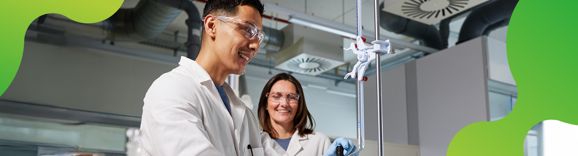 Eine junge Person führt eine Arbeit im Labor durch. Eine weitere Person schaut dabei zu. Beide lächeln und tragen Laborkittel, sowie Schutzbrillen.