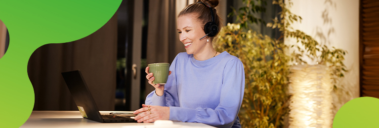 Eine junge Frau sitzt an einem Laptop. Sie lächelt und hält eine Tasse in der Hand. Sie trägt ein Headset und schaut auf den Bildschirm des Laptops. 