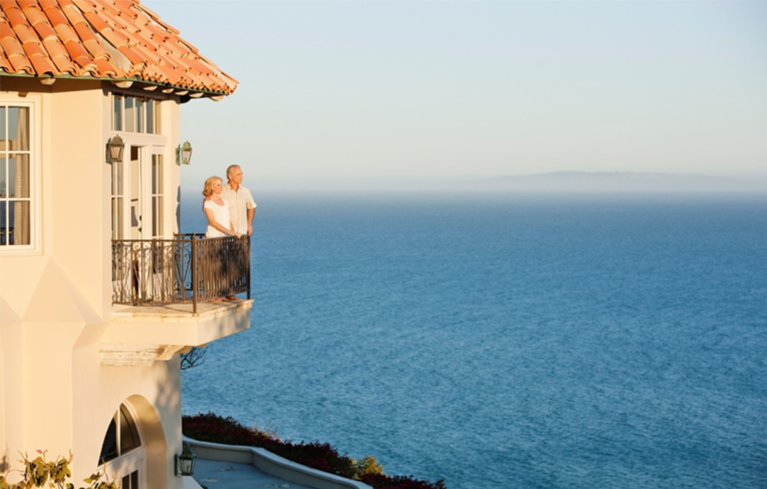 Ein Paar steht auf dem Balkon eines Hotels, schaut auf das blaue Meer und genießt seinen Urlaub trotz chronischer Erkrankung