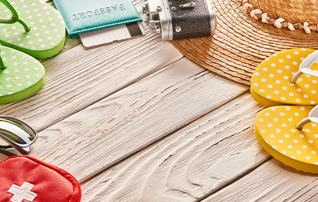 Verschiedene Reiseutensilien, wie Flip-Flops, Pass, Kamera, Sonnenbrille, Sonnenhut und Reiseapotheke liegen auf einem Holzboden