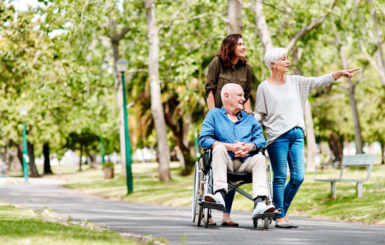 Eine Tochter geht mit ihrer Mutter und ihrem im Rollstuhl sitzenden Vater durch den Park