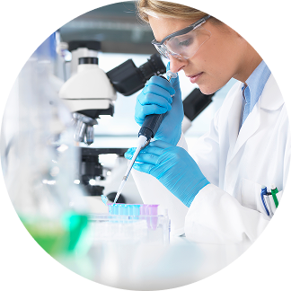 Eine Wissenschaftlerin im weißen Kittel und Schutzbrille entnimmt mit einer Pipette eine Probe und arbeitet an einem Mikroskop im Labor.