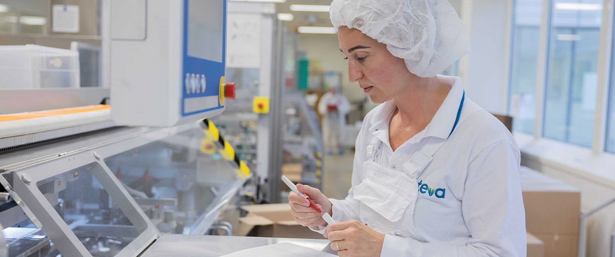 Frau in weißer Arbeitskleidung mit Haarnetz kontrolliert Verpackung an Produktionsmaschine im Teva Werk. 