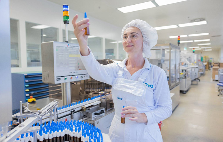 Frau in weißer Arbeitskleidung mit Haarnetz prüft braune Flasche an Abfüllanlage in der Teva Produktion. 