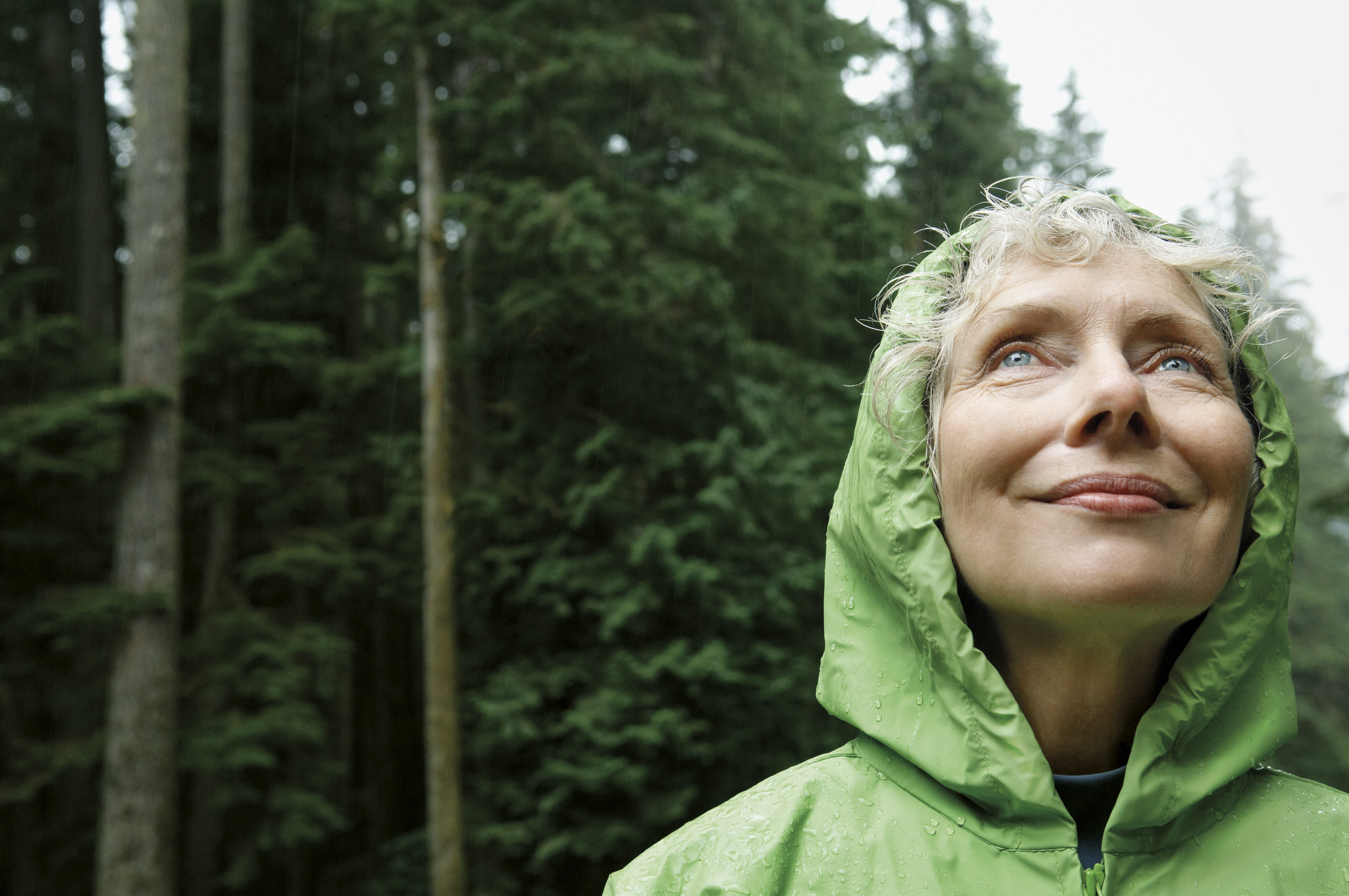 Ältere Frau in grüner Regenjacke genießt lächelnd die frische Luft im Wald