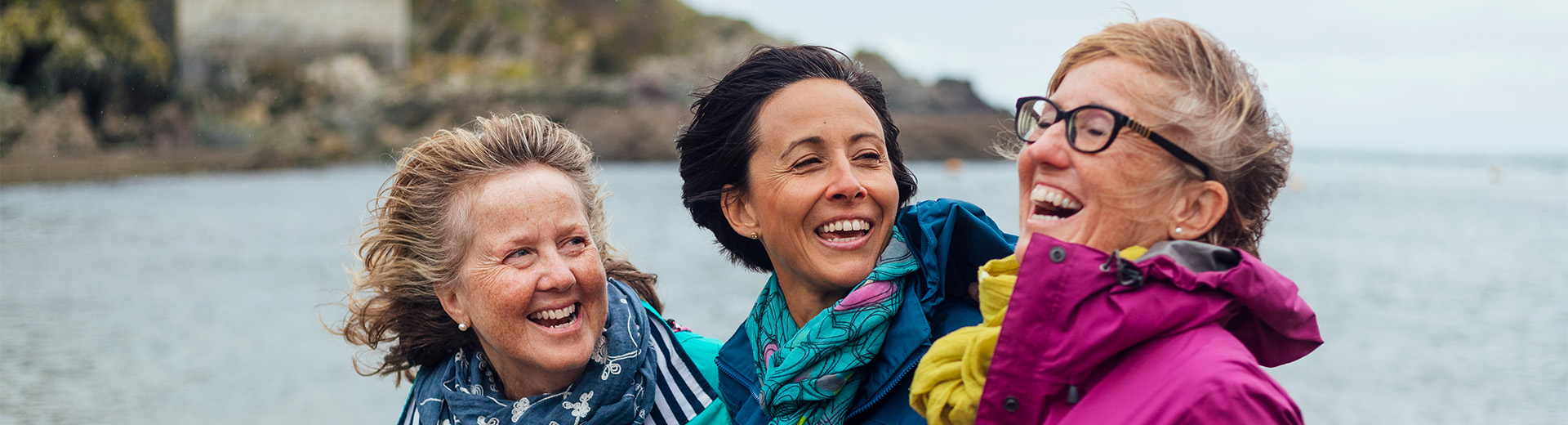 Drei Frauen mittleren Alters stehen an einem Strand in Outdoorjacken. Sie halten sich lachend im Arm.