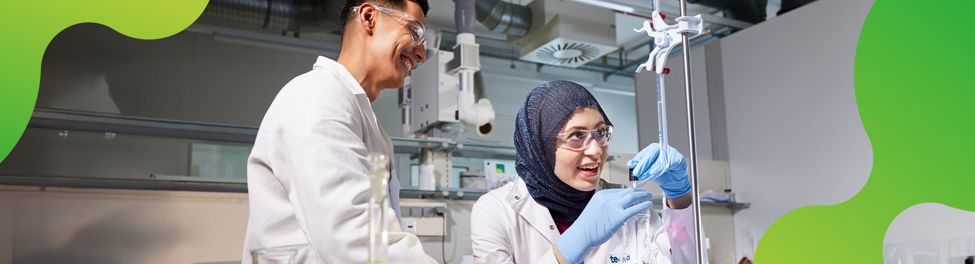 Eine junge Frau und ein junger Mann machen ein Experiment im Labor. Beide sehen begeistert aus und tragen Laborkittel, sowie Handschuhe und Schutzbrillen. 