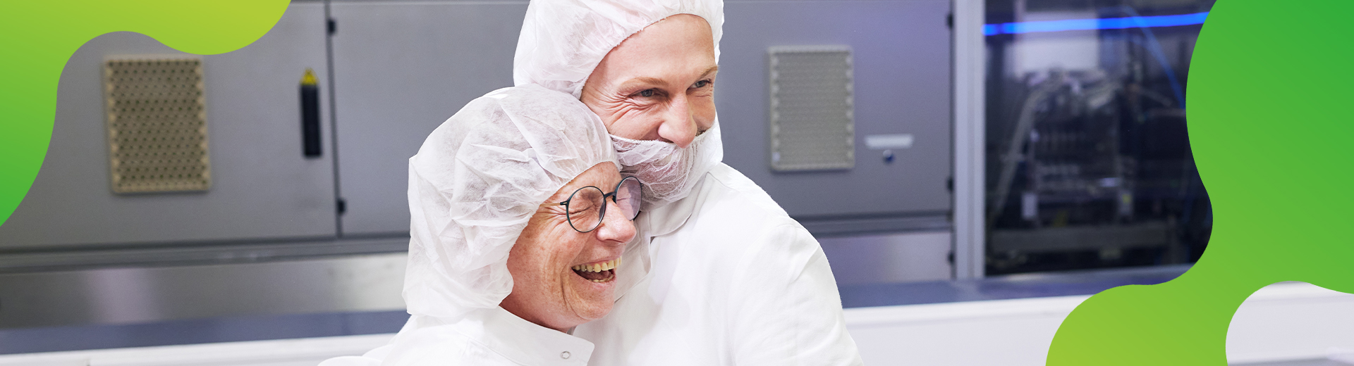 Zwei Personen in der Pharmaproduktion umarmen sich und lachen herzlich. Beide tragen Haarnetze und Laborkittel.