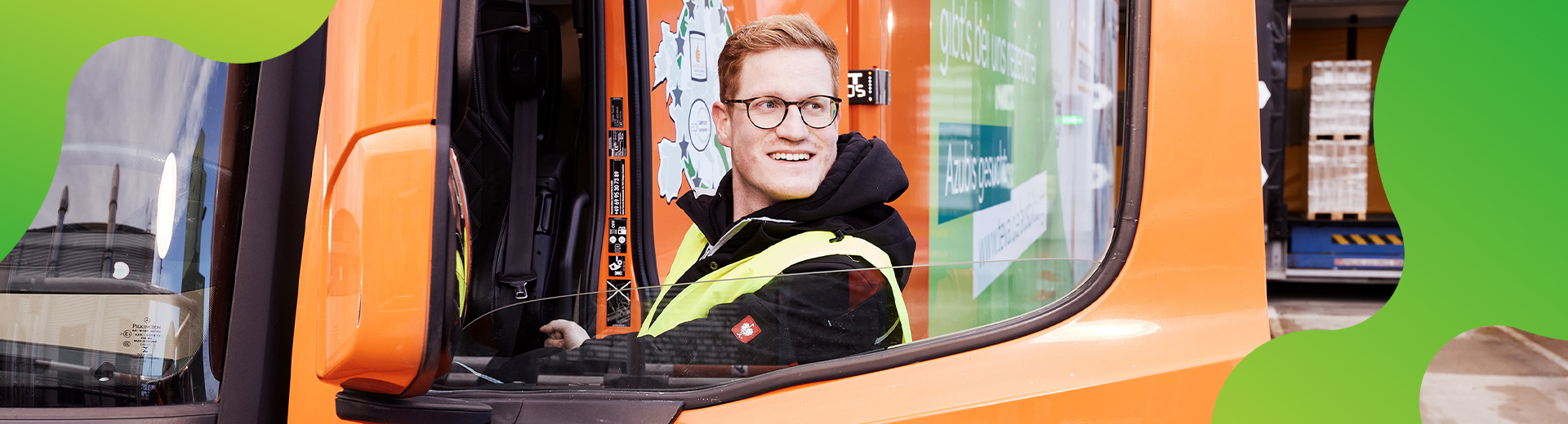 Ein junger Mann schaut durch ein geöffnetes Fenster eines orangenen LKWs er schaut lächelnd an der Kamera vorbei.