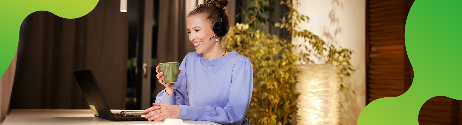 Eine junge Frau sitzt an einem Laptop. Sie lächelt und hält eine Tasse in der Hand. Sie trägt ein Headset und schaut auf den Bildschirm des Laptops.