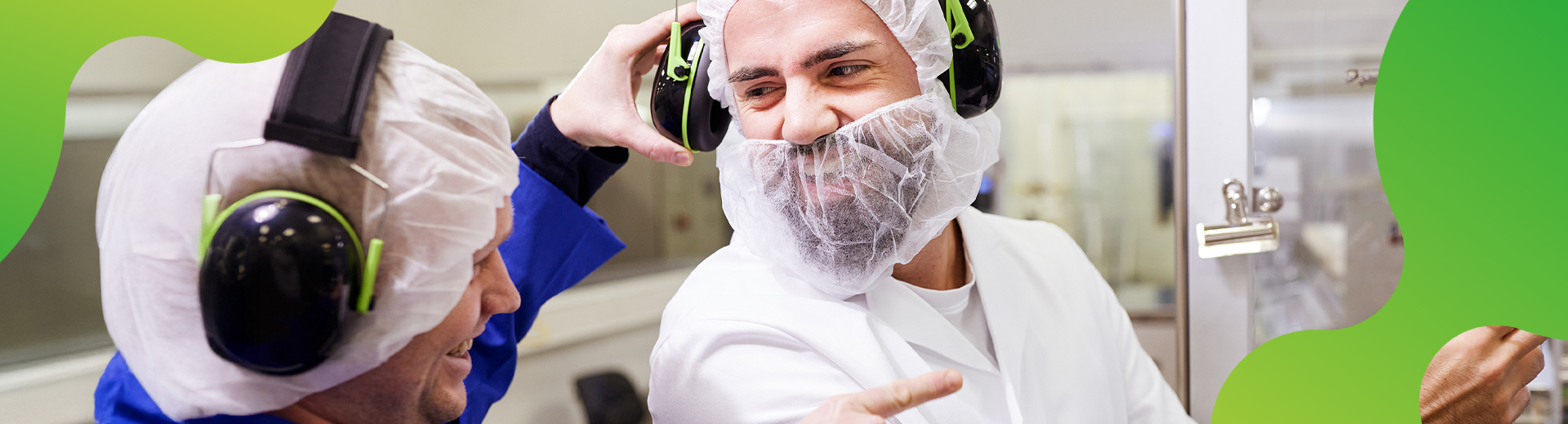 Eine Person in der Pharmaproduktion dreht sich zu einer anderen Person um. Beide lachen. Beide Personen tragen Haarnetze, sowie einen Gehörschutz.
