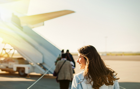 Von Migräne betroffen? 5 Tipps für den Urlaub Eine junge Frau mit langen braunen Haaren steigt in ein Flugzeug und schaut Richtung Sonne