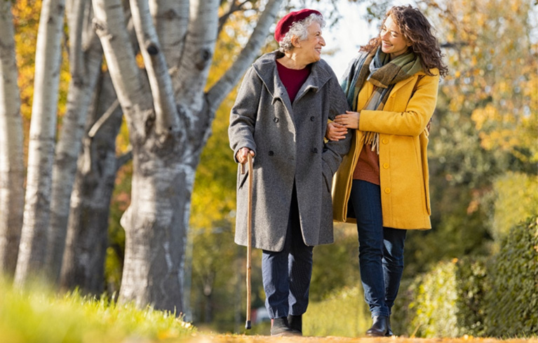 Eine Tochter geht Arm in Arm eingehakt mit ihrer pflegebedürftigen Mutter im Park spazieren